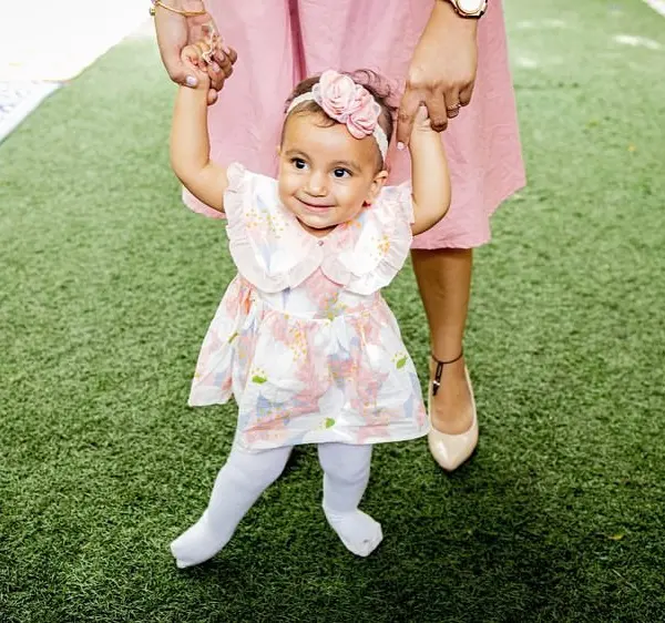 A baby girl in a floral dress and a pink headband taking steps on artificial grass, holding her mother's hands, at her first birthday party