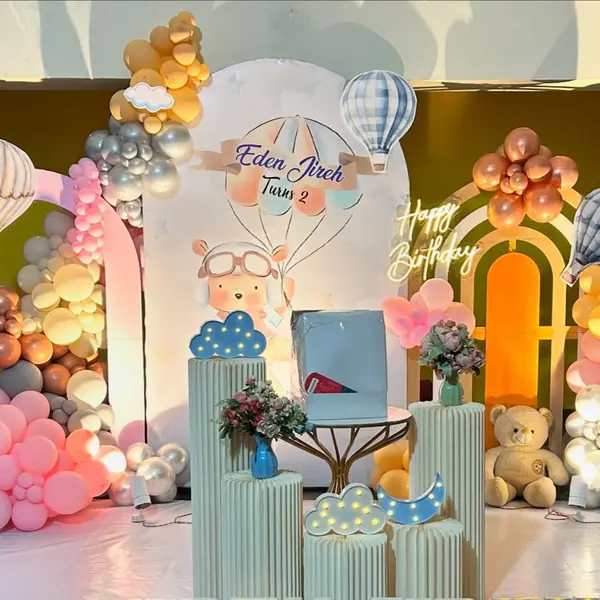 A second-birthday stage in a hall: three arched panels with a bear pilot under a hot-air balloon, balloon clouds in peach, blush, cream, silver and copper, hot-air-balloon cutouts, fluted plinths and two teddies