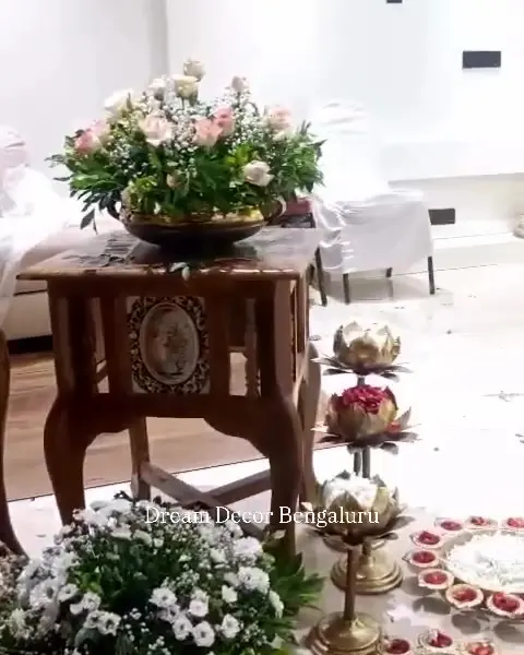 Lotus-shaped brass lamps on tiered stands, rings of small brass diyas with red petals and white flowers on the floor, and a carved table with a bowl of pink roses