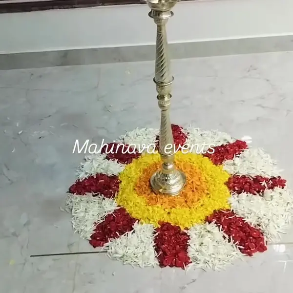 A round rangoli of white, red and yellow petals round a tall brass lamp, on a marble floor at a housewarming