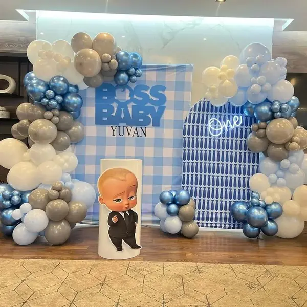 A Boss Baby first birthday: a blue gingham printed backdrop reading Boss Baby Yuvan, a second arched panel with a white One neon sign, a grey, white and blue balloon garland round both, and one Boss Baby cutout on a white plinth