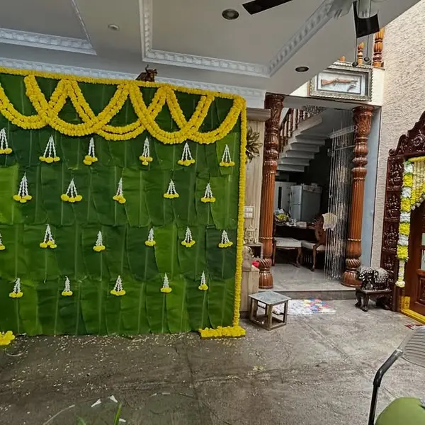 A wall of banana leaves about 8 feet wide in the hall of a house, with a scalloped swag of marigold along the top and small jasmine and marigold drops, beside a carved wooden door framed in flowers