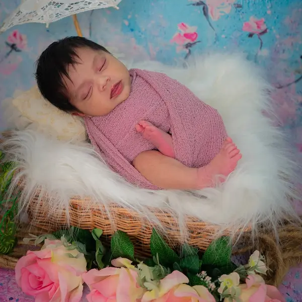 A newborn asleep in a wicker basket on white fur, wrapped in dusty-pink knit, against a painted floral backdrop with a lace parasol and pink roses