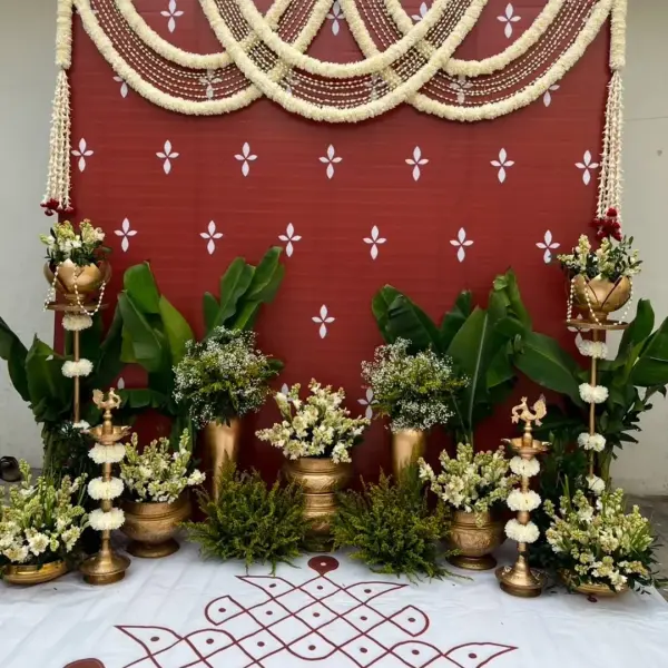 A red cloth backdrop with white motifs under scalloped garlands of white flowers, with banana plants, brass lamps hung with white flowers, pots of white flowers and a kolam on the floor sheet