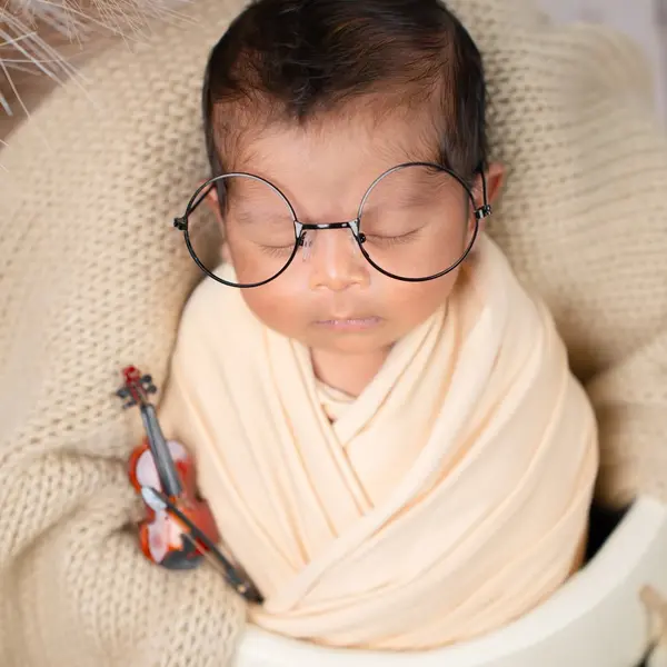 A newborn asleep in a cream wrap and round glasses with a toy violin, in a knitted bowl with pampas behind