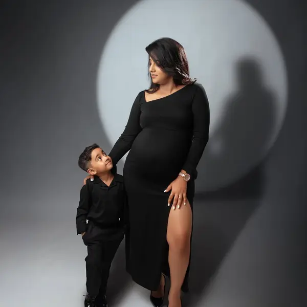 A pregnant woman in a black long-sleeved dress stands under a circle of light on a grey backdrop while a small boy in a black suit looks up at her