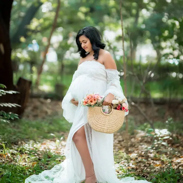 A pregnant woman in a white off-shoulder tulle gown stands among fallen leaves holding a straw basket of flowers, trees behind her