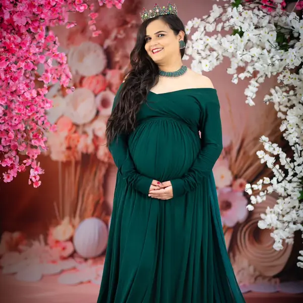A pregnant woman in a dark green long-sleeved gown and a crown stands against a wall of pink and white blossom