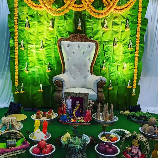 A seemantham stage: a banana-leaf wall with marigold swags and small jasmine and marigold drops, a white and carved-wood throne, and in front rows of plates of fruit, sweets and coconuts, a pineapple, a framed set of dolls and a small doll in a cart