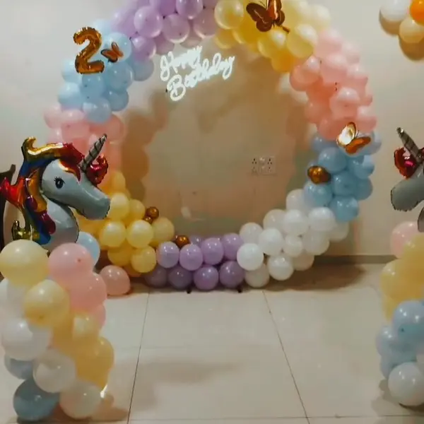A full pastel balloon ring in lilac, yellow, peach, blue and white with gold butterflies, a white Happy Birthday neon sign, a gold 2 and a unicorn foil balloon on a balloon column, in a hall