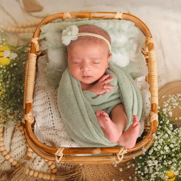 A newborn asleep in a cane basket, wrapped in sage-green knit with a flower headband, feet drawn up and one hand by the chin, among baby's breath and beads