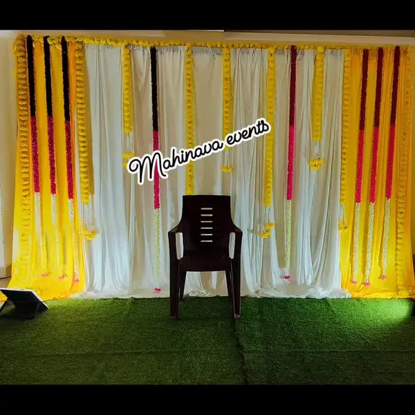 A wide backdrop of yellow and white drapes hung with long strings of marigold and red flowers, on an artificial grass mat with a plastic chair in front
