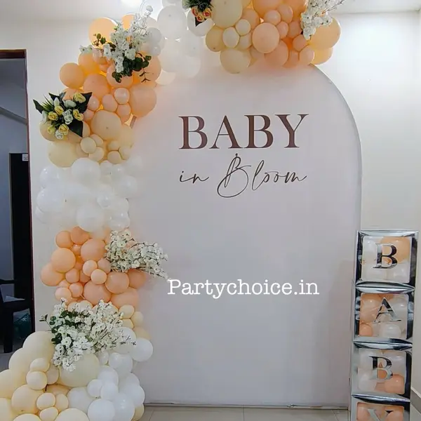 At home: a tall cream arched panel reading BABY in Bloom, a peach, cream and white balloon garland with white blossom sprays, and four silver BABY boxes