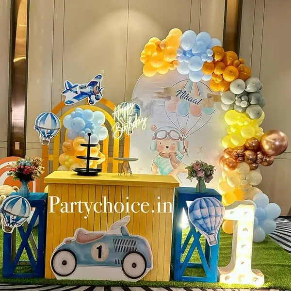 A first birthday in a hotel hall: a round print of a bear pilot under balloons, a yellow and blue garland, a neon sign, a yellow cake table with a race-car front, plane and hot-air-balloon cutouts, blue plinths, a lit 1 and a grass carpet