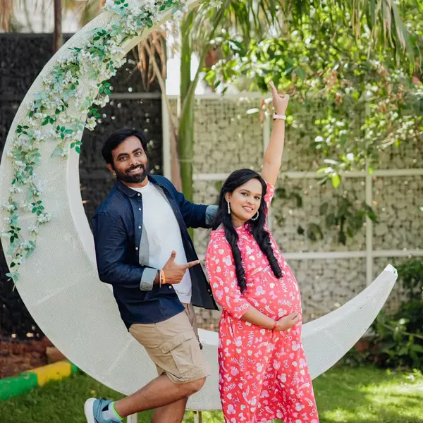 A pregnant woman in a pink print dress raises one arm beside a white crescent-moon swing wrapped in leaves, her husband in a denim jacket leaning on it, on a green lawn with palms
