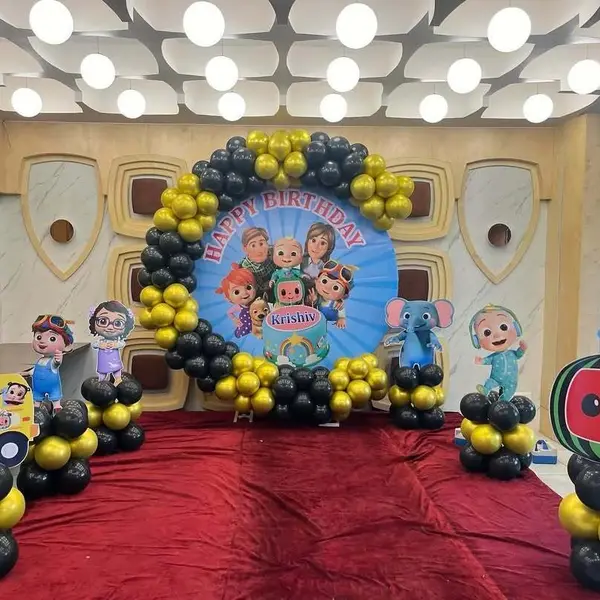 A Cocomelon setup on a red carpet: a round print reading Happy Birthday Krishiv in a black and yellow balloon ring, and six character cutouts on balloon columns, including the school bus and the watermelon