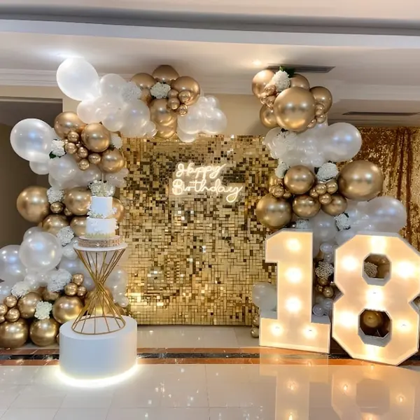 An 18th birthday in a hall: a gold sequin wall with a Happy Birthday neon sign, a white and gold balloon garland with white flowers, a two-tier cake on a gold stand and a lit 18 about 3 ft tall