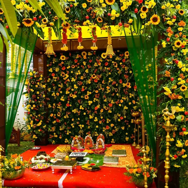 A mandap in a home garden with a yellow roof and hanging bells, a wall of sunflowers, marigold and gerbera behind, green curtains, tall brass lamps, kalashas with garlands and offerings on a red carpet