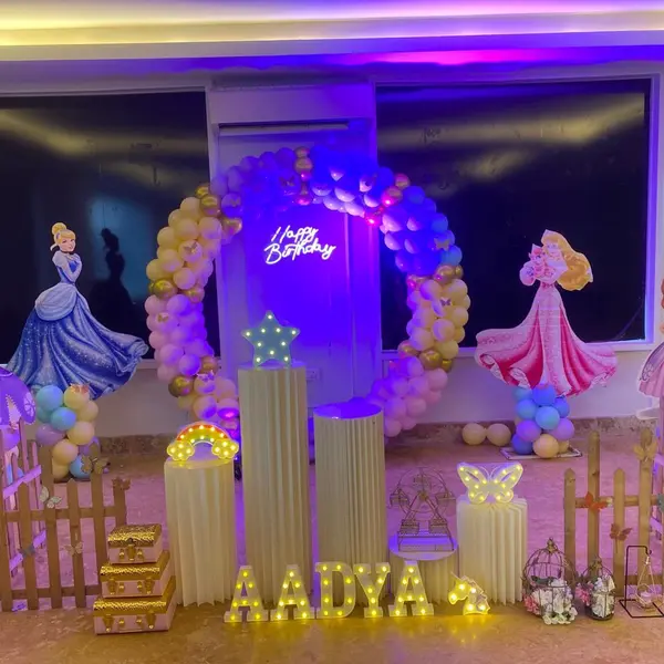 A hall setup: a balloon ring with a Happy Birthday neon sign, four life-size princess cutouts, wooden picket fences, five fluted plinths with a lit star, rainbow and butterfly, trunks, birdcages and the name AADYA in lit letters