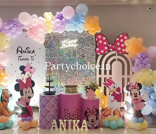 A Minnie Mouse setup indoors: a silver shimmer wall with a neon sign, a pastel balloon arch, character cutouts including Minnie, Daisy and Pluto, three pink plinths and the name ANIKA in five lit letters