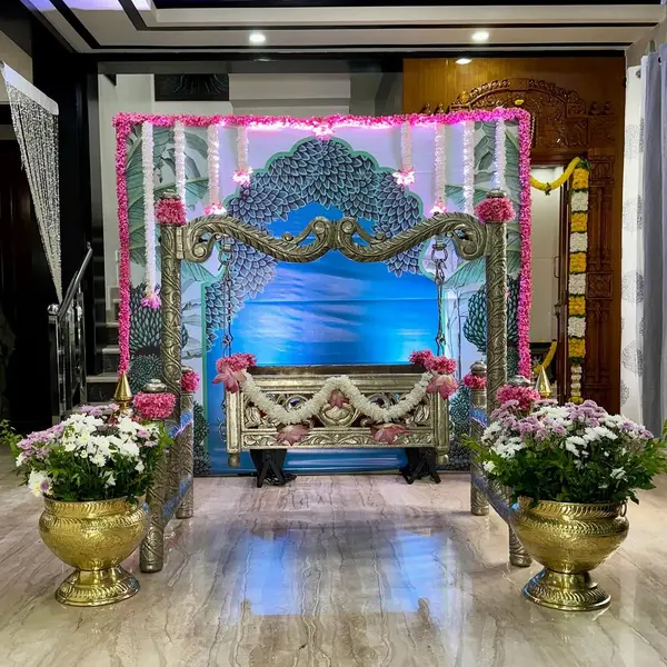 A carved brass swing on chains wrapped in pink flowers, a white flower garland across its seat, a pink flower frame round a printed peacock-feather backdrop, and two brass urlis of white and lilac chrysanthemums, on a marble floor
