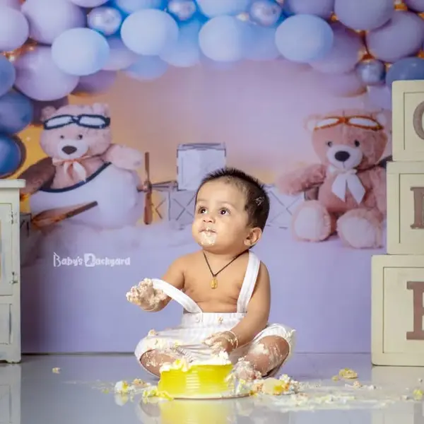 A baby sitting in a smashed yellow cake in a studio set with lilac and navy balloons, teddy bears in pilot goggles and wooden ONE blocks