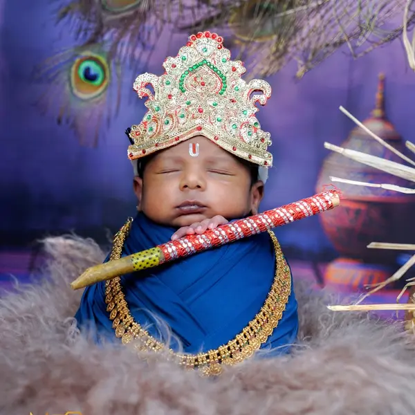 A newborn asleep in a jewelled Krishna crown and a blue wrap with a gold necklace and a flute, on grey fur under peacock feathers