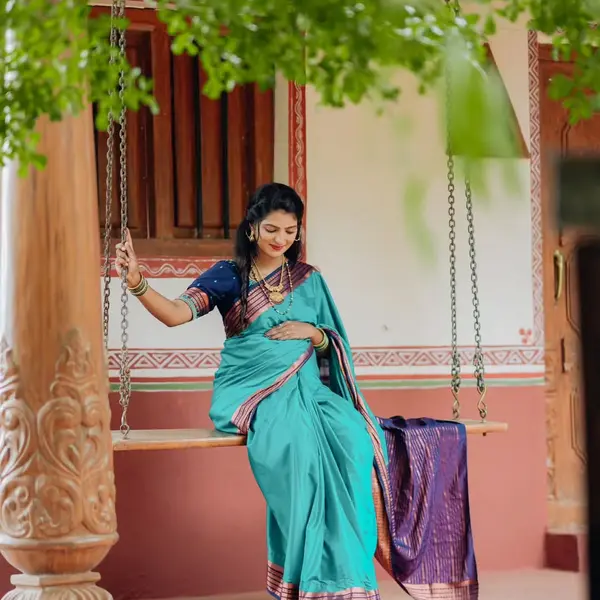 A pregnant woman in a teal saree with flowers in her hair sits on a carved wooden swing in front of a pillared red and white house