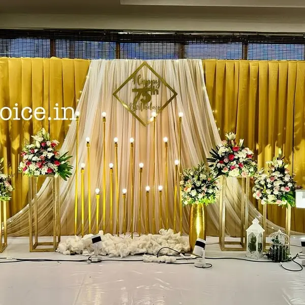 A 75th birthday stage: gold pleated curtains with a cream drape in the middle, a gold Happy 75th Birthday sign, rows of candle poles, flower stands, lit 75 numbers and a white floor sheet