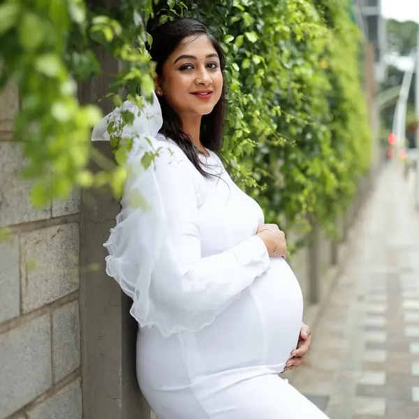 A pregnant woman in a white fitted gown with sheer ruffled sleeves leans on a stone wall beside a clipped hedge on a paved path