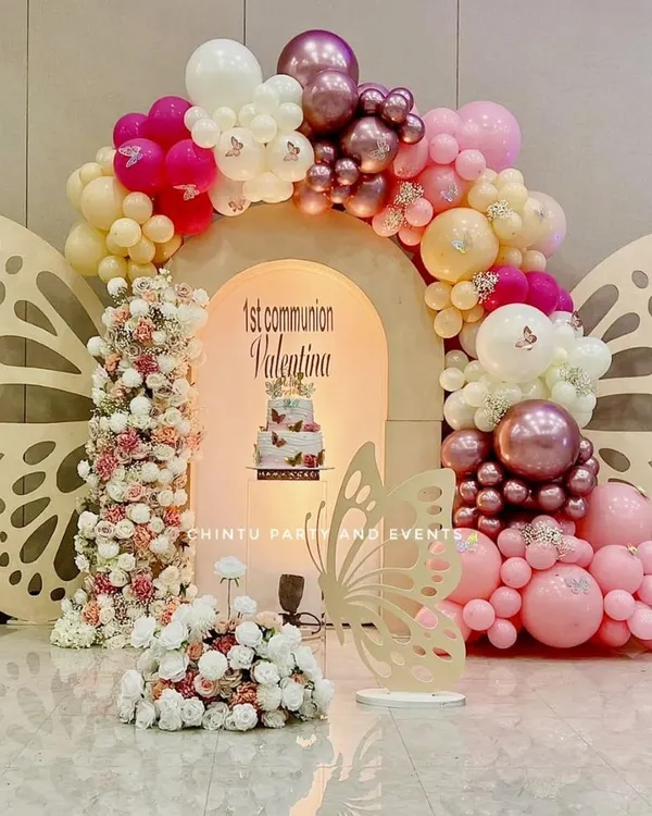 A beautiful butterfly-themed First Holy Communion decoration. This setup features large butterfly wing cutouts, a pink and white balloon arch, and floral arrangements.