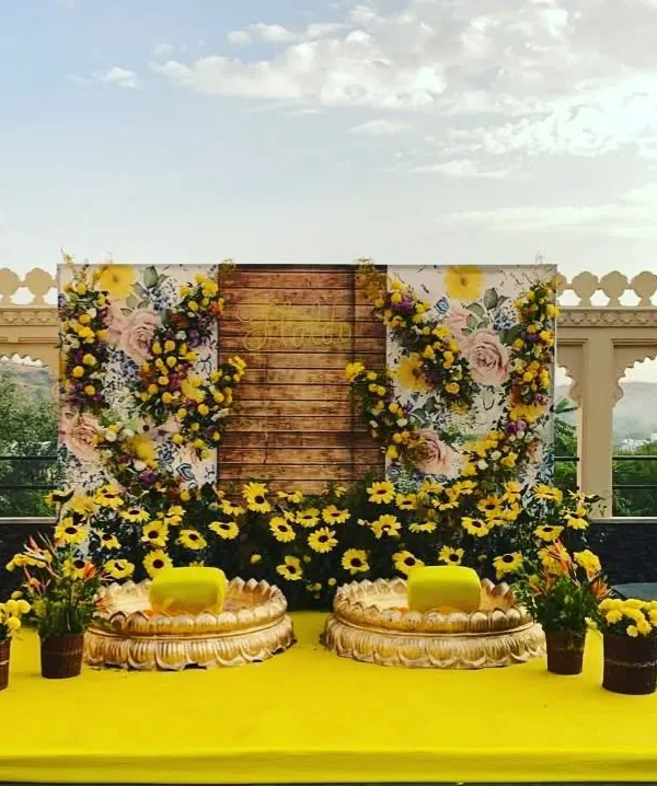 A stunning Haldi stage setup against an open sky. The backdrop combines a rustic wooden panel with a floral print and is adorned with sunflowers and marigolds for a fresh, picturesque look.
