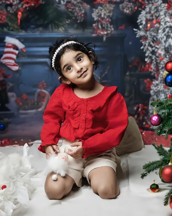 The magic of Christmas. A little girl in a festive red outfit sits by the tree, her eyes sparkling with holiday wonder in this cozy studio setup.