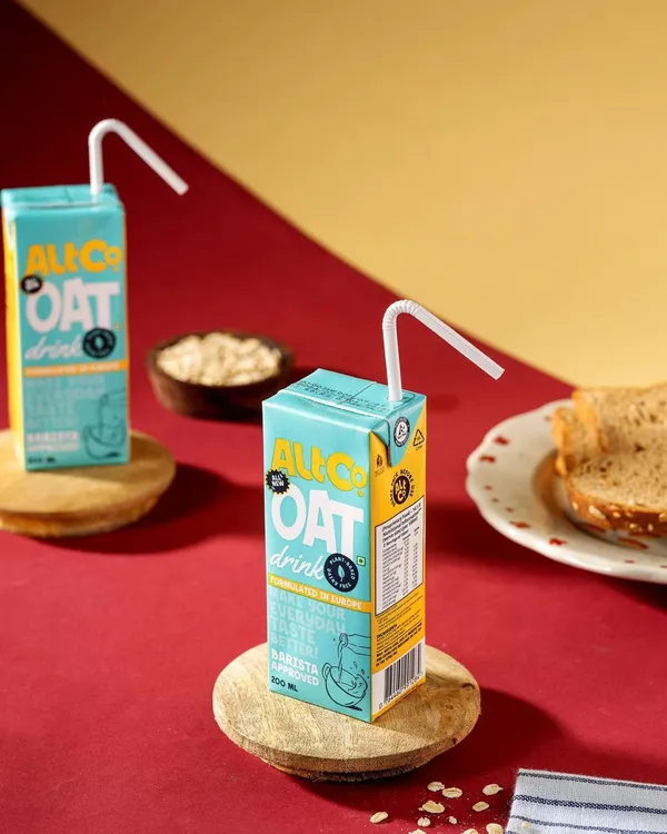 A graphic and colorful studio shot of Alt Co. Oat Drink. The bold red and yellow background with wooden coasters creates a modern, pop-art feel that is eye-catching and playful.