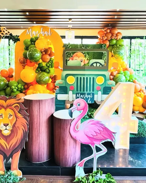 A vibrant and immersive jungle safari theme for a fourth birthday, featuring life-sized lion and giraffe props, a custom jeep cutout for photos, and a large light-up number '4'. The scene is framed by a lush balloon garland in shades of green, orange, and brown, creating a true wild adventure.