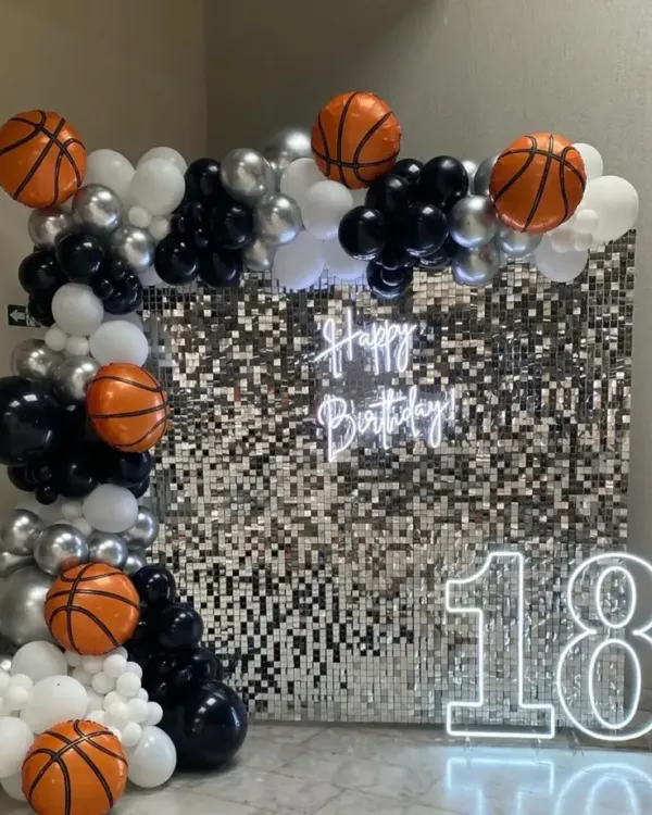 A modern and stylish 18th birthday setup. This features a shimmering silver sequin wall, a custom "Happy Birthday" neon sign, and a large "18" marquee number, all framed by a balloon arch in black, white, silver, and basketball-themed balloons.