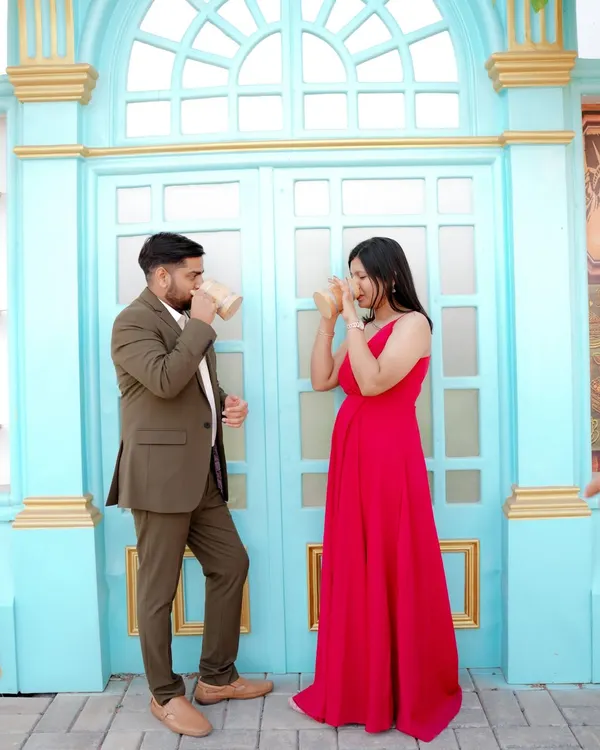 A playful moment sharing a drink against a vibrant blue door, reminiscent of a European street. The woman's bright red dress pops against the cool tones, creating a lively and stylish pre-wedding photo.
