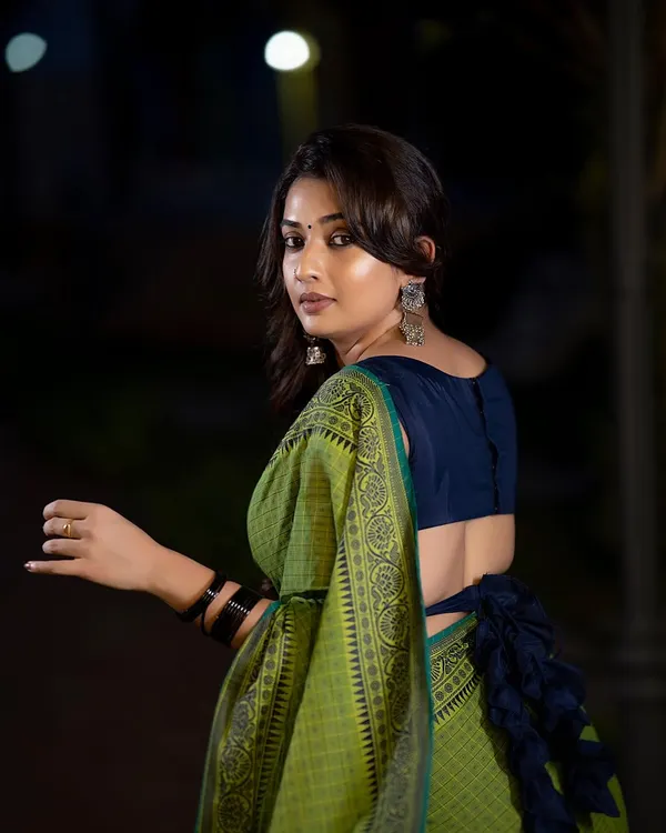A beautiful outdoor portrait at dusk. The low light and backlighting create a soft glow around the subject, highlighting the elegant drape of the green and blue saree.