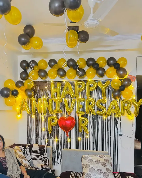 A 'Happy Anniversary' decoration in a living room, using black and gold balloons, a shimmer curtain, and fairy lights.