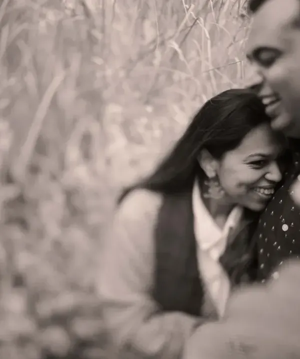 A soft, grainy black and white photo that feels incredibly intimate and timeless. The focus is entirely on the couple's happy embrace, surrounded by tall grass.