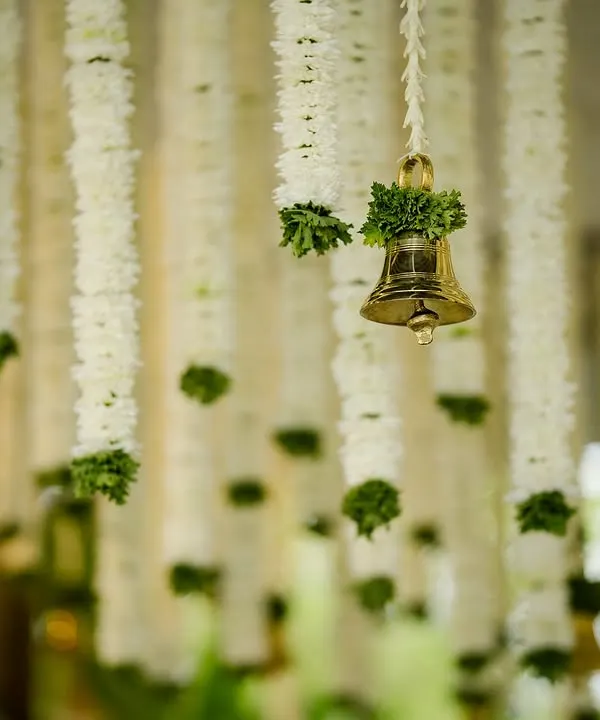 A close-up of hanging floral decor, featuring strings of white chrysanthemums and a single brass temple bell. This simple detail adds both visual beauty and a touch of serene sound to the ambiance.