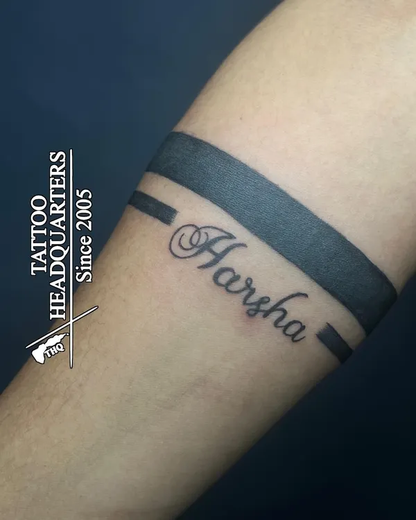 A classic armband tattoo personalized with the name "Harsha" in a beautiful cursive script, integrated into the solid band design.