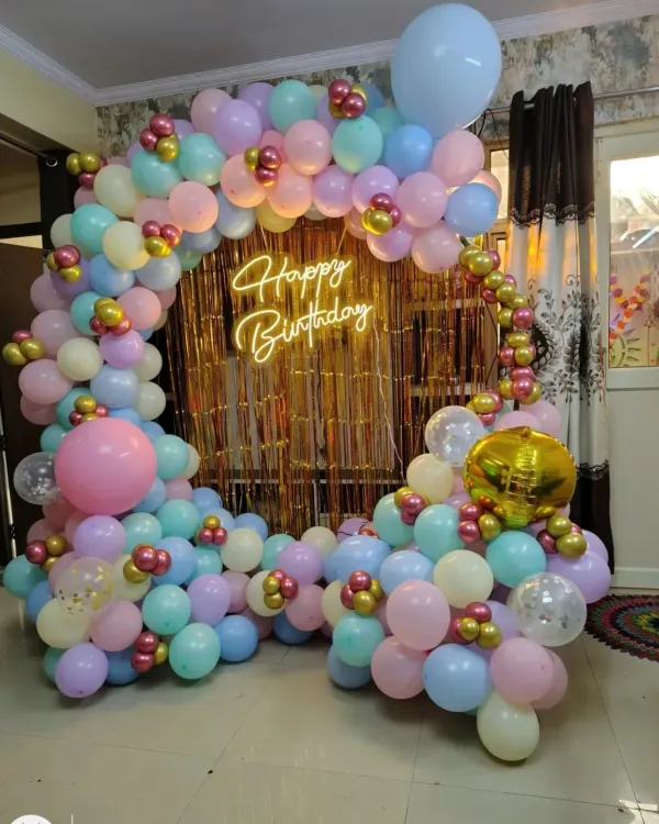 A colorful pastel balloon ring with a gold fringe backdrop and a "Happy Birthday" neon sign, perfect for a cheerful home party.