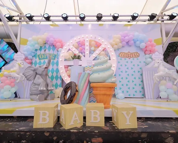 A dreamy pastel carnival stage setup for a first birthday. The scene includes a lit Ferris wheel, giant ice cream cone and donut props, and adorable animal figures, all in front of a custom-designed geometric backdrop.