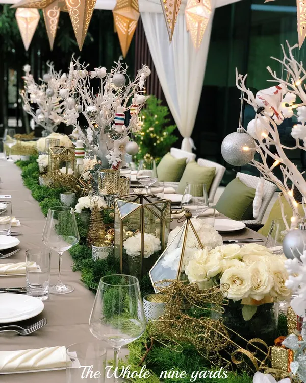 A beautifully set table for a Christmas Eve dinner. The decor combines a natural feel with festive elegance, using a green runner, white florals, and sparkling gold and silver accents.