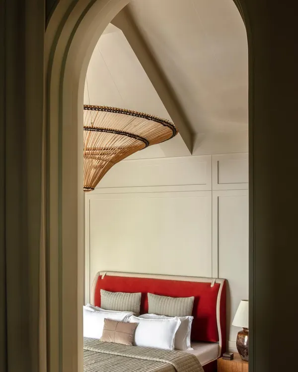 An arched doorway frames the master bedroom in our 'Summer Haus' project, creating a sense of theatrical entry. The view reveals a bold red headboard and a striking rattan light fixture, hinting at the eclectic and cosmopolitan design within.