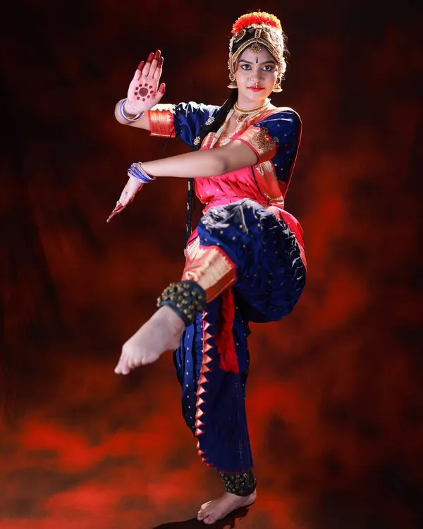 A dynamic full-body shot of a Bharatanatyam dancer in mid-pose. The dramatic red background and studio lighting make the dancer's form and colorful costume stand out.