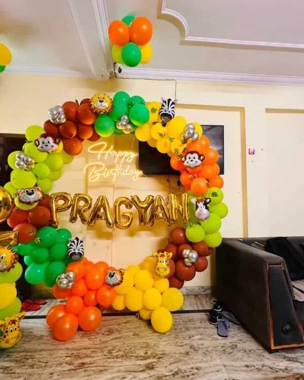 A jungle-themed decoration for a second birthday party, set up right in the living room. The design is built around a circular frame, featuring a colorful balloon garland, cute animal face cutouts, and personalized foil balloons for the birthday boy's name.
