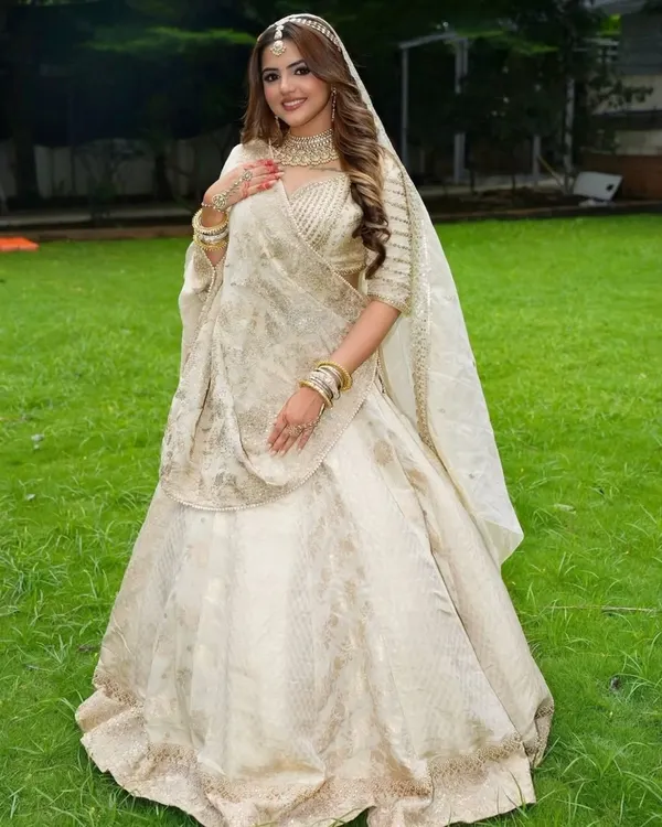 A full-length view of a bride in a magnificent ivory and gold lehenga. The look is completed with a matching choker, bangles, and a delicate matha patti for a truly royal feel.