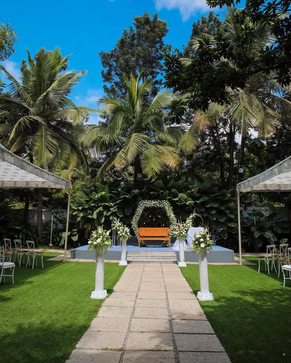 This is the heart of The Farmhouse Collective, set for an intimate wedding. The stone pathway leads to a beautiful stage, surrounded by lush lawns and tropical greenery, creating a perfect natural aisle under a brilliant blue sky.